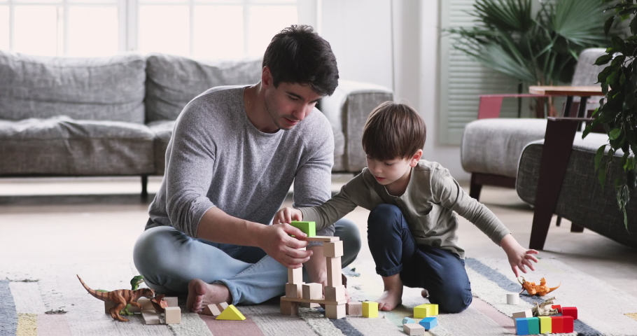 Happy young adult parent dad and child son play toys sit on floor carpet at home. Caring father having fun help cute kid build tower of wooden blocks. enjoy game activity give high five in living room | Shutterstock HD Video #1047050953