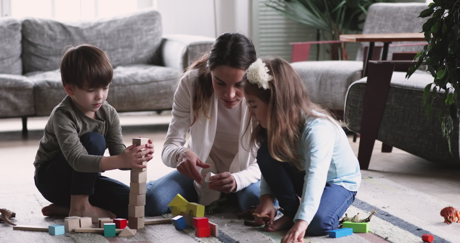 Happy young adult mother nanny playing with two cute small kids, having fun sit on floor carpet at home. Playful mum helping preschool children building tower of wooden blocks in living room Royalty-Free Stock Footage #1047050968