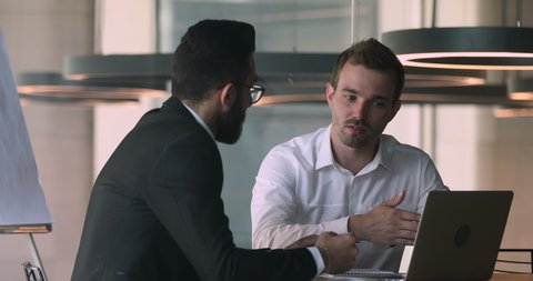 Happy young caucasian businessman discussing online project presentation with smart arabian male partner in office. Confident salesman financial advisor explaining deal details to arabic client.