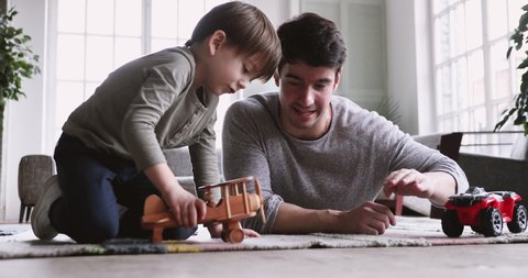 Young father and cute small kid son playing funny racing game relax on carpet floor at home. Happy male family adult dad and little child boy having fun with toy car plane engaged in leisure activity