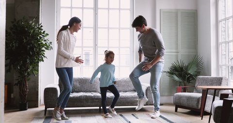 Happy playful young family couple dancing with cute active preschool  small child daughter in living room interior together. Adult parents mum dad and kid girl pretending running having fun at home