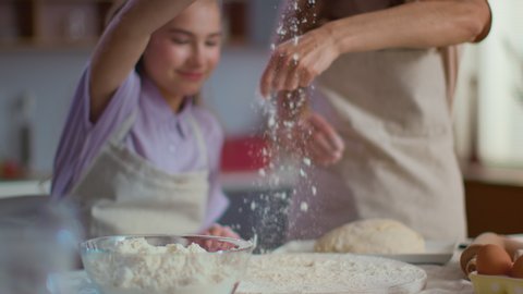 Closeup of woman and girl sprinkling flour on table at kitchen in slow motion. Mother and daughter cooking dough for cookies on modern kitchen. Family spending time together at home