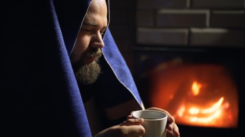 Man with beard covered with blanket drinks hot tea from a cup near fireplace. Cozy hygge scene in the night in a countryside house. 