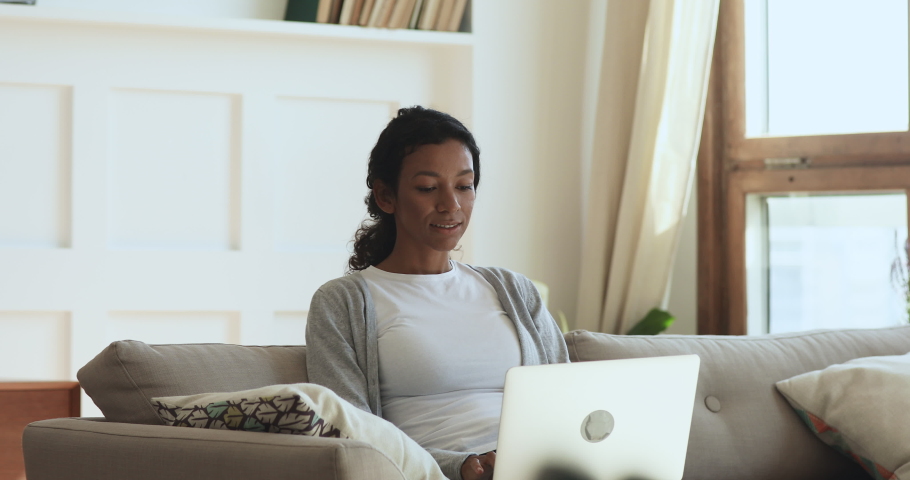 Happy millennial biracial girl relaxing at home, using computer apps, making purchases in online store. Skilled woman working on laptop remotely. Smart student doing homework alone in living room. Royalty-Free Stock Footage #1047056821