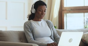 Smiling young african american woman wearing wireless headphones, holding video call with relatives using computer. Happy millennial biracial girl chatting online with friends, sitting on couch.