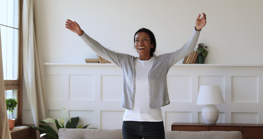 Overjoyed young african woman feeling joyful, thanking god for good luck. Thankful mixed race girl enjoying freedom alone at home. Excited homeowner spinning with stretched arms, real estate concept. Royalty-Free Stock Footage #1047056845