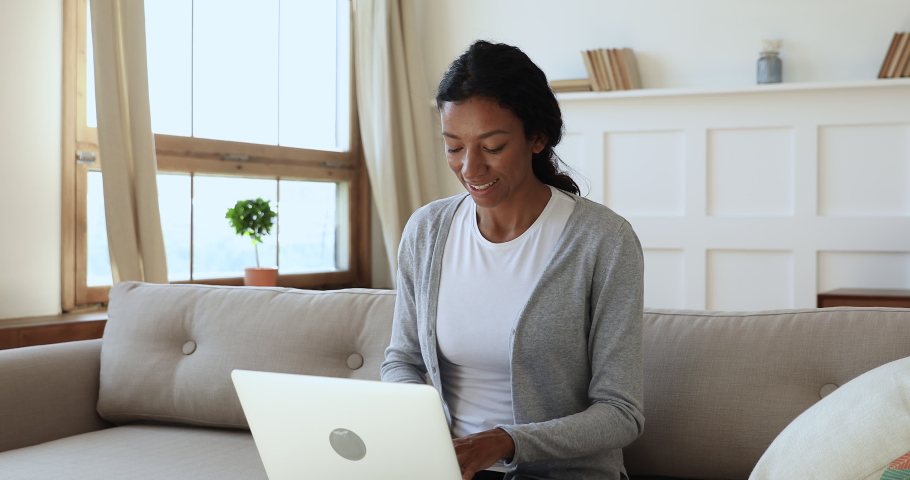 Focused young african ethnicity woman sitting on couch, typing message on computer, happy chatting with friends. Smiling millennial businesswoman freelancer satisfied with client reply on offer. | Shutterstock HD Video #1047056854