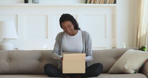 Curious millennial mixed race girl sitting on sofa, unpacking cardboard parcel, surprised by fast delivery service. Interested young african american woman unboxing purchase from internet store.