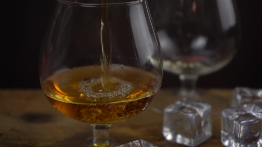 Cognac \ whiskey pouring into glass, golden color strong spirit alcohol in cognac glasses on dark black background, slow motion footage | Shutterstock HD Video #1047057100