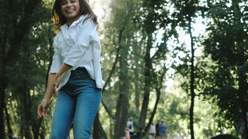 Spring photo shoot for a cute girl in white shirt and jeans. Cheerful young woman with long hair and white manicure is dancing at the camera among the trees in the park on sunny day. Video for memory.