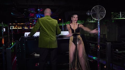 Long shot of young Caucasian woman in seductive dress dancing next to elderly male DJ in night club. Mature man in green jacket mixing music on controller as professional PJ moving on stage.