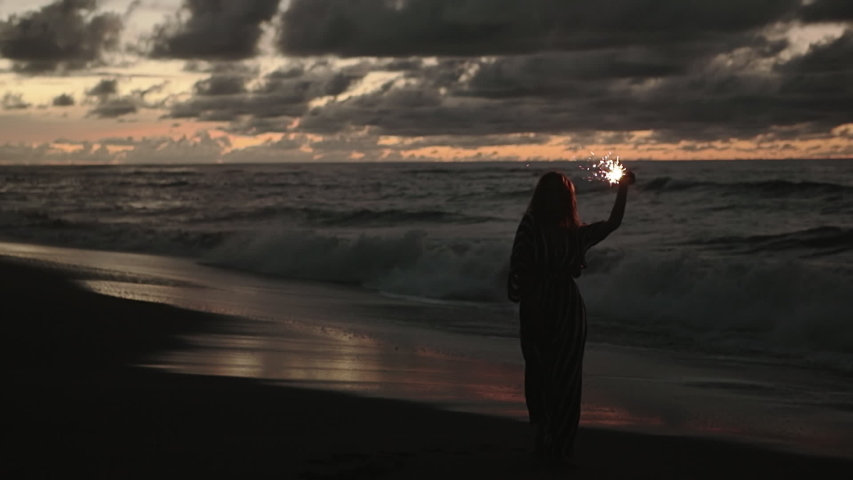 Close up portrait of young beautiful woman at sunset on beach. Pretty girl dancing in with sparkler at sunset in slow motion. Holiday with fireworks. City people travel. Business woman at vacation.