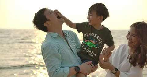 Happy parents kissing their son in both cheeks. Young asian father and mother are holding their cute child in their hands and kissing him from both sides on the beach at sunset.