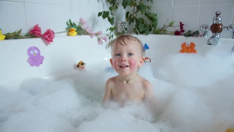 Little blue-eyed child in bath filled with foam, playing with toys and smiles, mother's hands soap the baby's head with baby shampoo