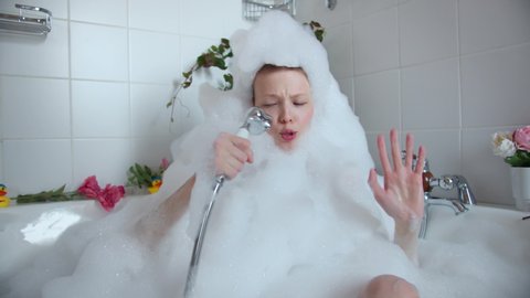 Funny girl sits in bath filled with foam, holds shower singing songs. Girl in foam from head to toe in the bath fooling around