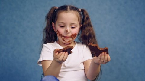 Dirty face of a girl eating chocolate. Two pieces of sweet chocolate in child's hands. Portrait of a little kid with smeared face isolated in studio.