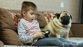 Little boy plays in video game with joystick, pug dog with child on the couch at home on vacations