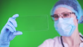 green background. close-up, doctor dressed in medical cap, mask, blue medical gloves, holds a glass card and clicks on it. its possible to place advertisement, text or video on card.