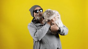 Stock footage of a cool senior bearded man in hoodie, hat, sunglasses and headphones talking to a lovely pet bunny in his hands. Cutout on yellow.