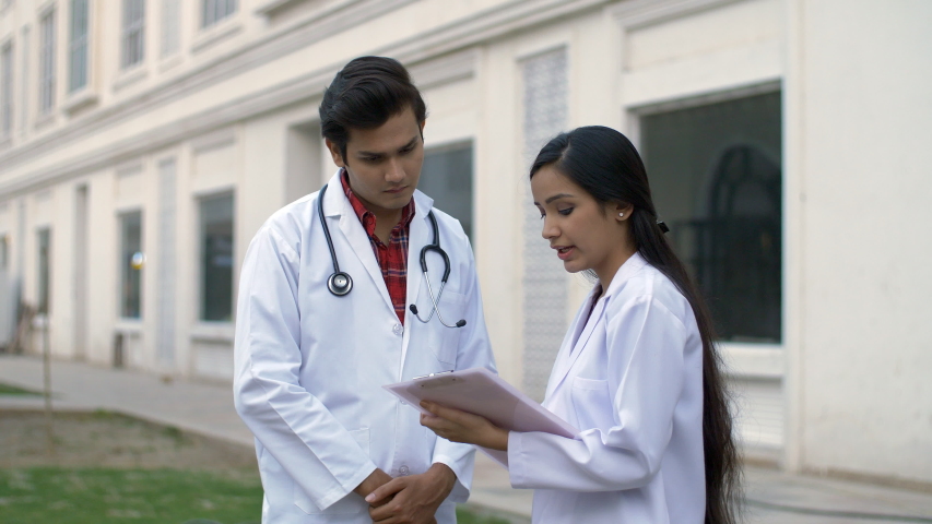 Two young doctors discussing the disease history of a patient outside the hospital. Indian medical professionals wearing white coat consulting on patient's medical records while standing in a hospi... | Shutterstock HD Video #1047111739