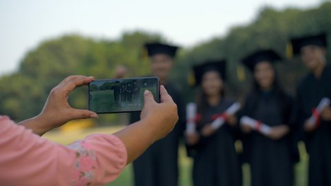 Pan shot - Woman hands clicking pictures of college pupils on graduation day. Happy group of young teens posing for the photos while holding their degrees in graduation gowns - convocation ceremony