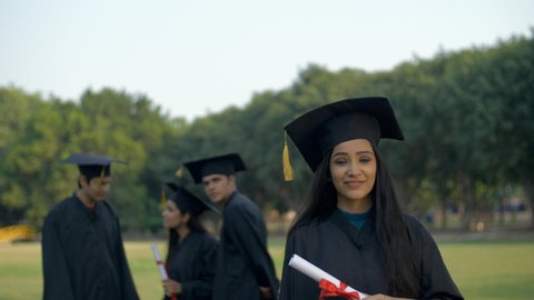 Attractive Indian graduate posing towards the camera with a big smile on her face. Young graduated girl holding her graduation degree in pride while wearing the academic black gown and cap - convoc...