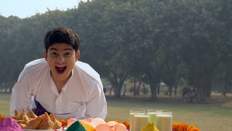 Pan shot of a cute Indian guy is really excited to celebrate the joyful Holi festival. Beautifully decorated festive table with colorful Gulal, Indian sweet Gujiya, Samosa, Kesar Thandai, water bom...