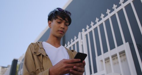 Front low angle view of a fashionable mixed race transgender adult hanging out on a summer day in the city, standing in the street and using his smartphone in slow motion.
