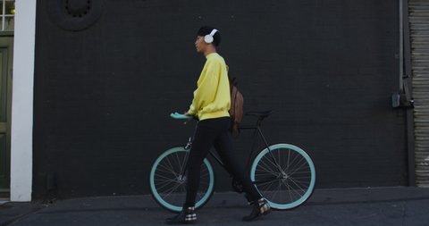 Side view of a fashionable mixed race transgender adult hanging out on a summer day in the city, wearing headphones, walking and wheeling his bicycle during a sunny day in slow motion.
