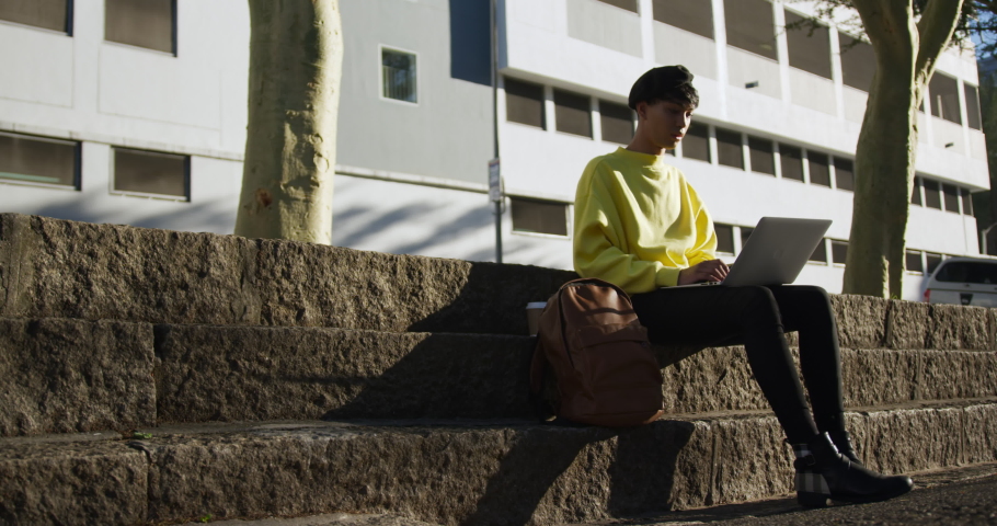 Side view of fashionable mixed race transgender adult hanging out on a summer day in the city, sitting in the street, sitting on the stairs in the street and using his laptop in slow motion.