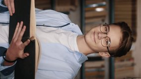 Vertical video of a happy teen boy in glasses reading a book, smiling looking at the camera while standing between rows of bookshelves in the library - Powered by Shutterstock - Get 15% off with code: PIKWIZARD15