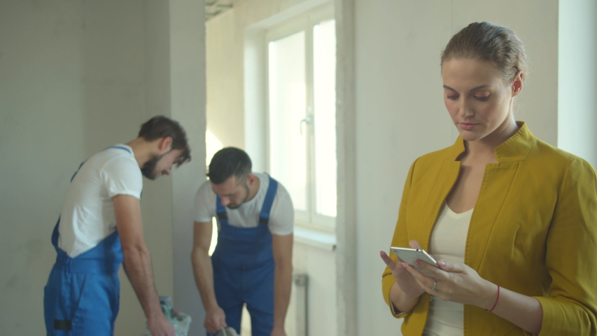 Woman takes a photo on phone, builders work in the flat