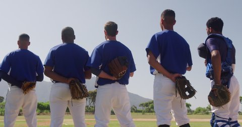 Rear view of a multi-ethnic team of male baseball players, preparing before a game, standing in a row, holding their caps on their chests, listening to national anthem on a sunny day in slow motion