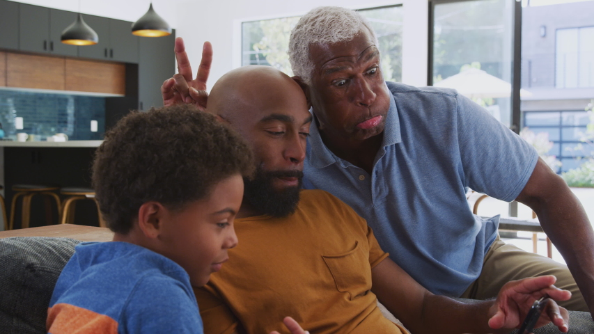 Multi-generation male african american family sitting on sofa at home pulling faces as they pose for selfie on mobile phone - shot in slow motion Royalty-Free Stock Footage #1047139657