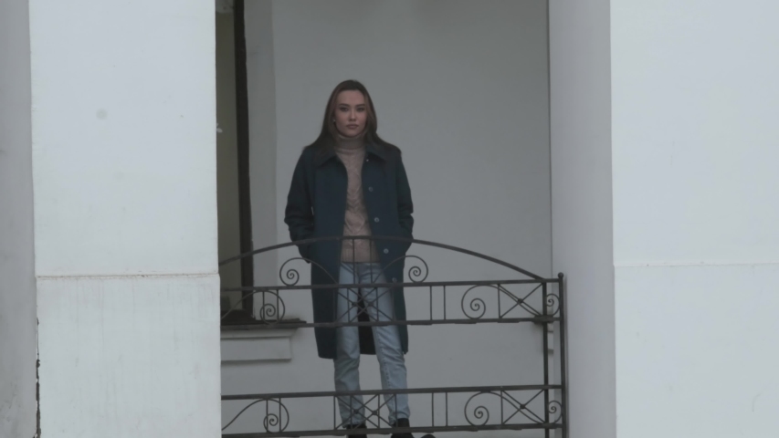 medium shot of stylish asian girl in blue coat and sweater with long hair standing outdoors between two white columns. cars and transport pass in front of her. Urban portrait of young female at street