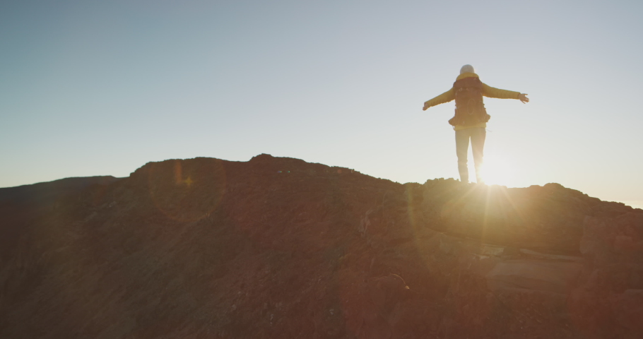 Young woman standing atop a mountain holding her arms up towards the sky enjoying the sunset outdoors, young hiker finishing her trek at sunset