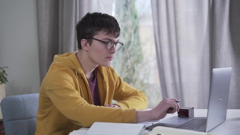 Camera moving from left to right around young Caucasian boy in eyeglasses writing and using laptop. Intelligent nerd student doing homework at the table. Education, intelligence, lifestyle.