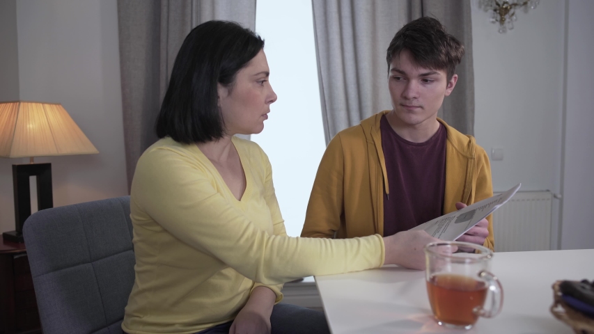 Portrait of strict dissatisfied Caucasian mother sitting with teenage son and checking list with his bad grades. Angry woman singing and looking at lazy college student. Failure, lifestyle, education. Royalty-Free Stock Footage #1047167218