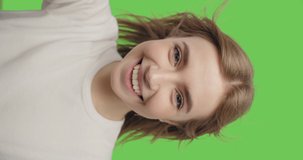 Young smiling hipster blond woman in white t-shirt posing over green screen background. Girl taking selfie self portrait photos on smartphone. Vertical 4k raw video footage slow motion 60 fps