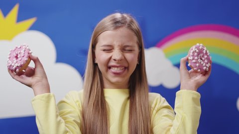Cute adolescent school girl plays with sweet donuts covering her eyes doing happy fun face expressions on background. Beautiful rainbow sunny decoration.