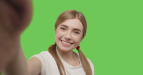 Young smiling hipster blond woman in white t-shirt posing over green screen background. Girl taking selfie self portrait photos on smartphone. Female model showing positive face emotions