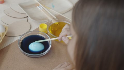 High angle shot of little girl dipping egg in blue food coloring in glass bowl and using wooden stick to turn it while preparing Easter decorations