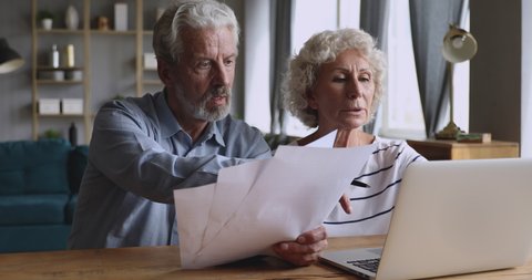 Focused middle aged man holding paper documents, helping pleasant attractive mature wife filling information in online banking app on computer. Serious older couple managing family budget at home.
