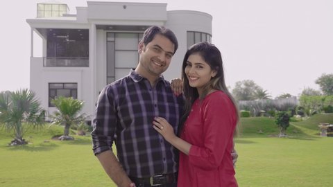 A wide shot of happy married couple standing outside luxurious house looking at the camera with a smile on their face. Attractive and smiling Husband and wife in front of newly constructed bungalow.