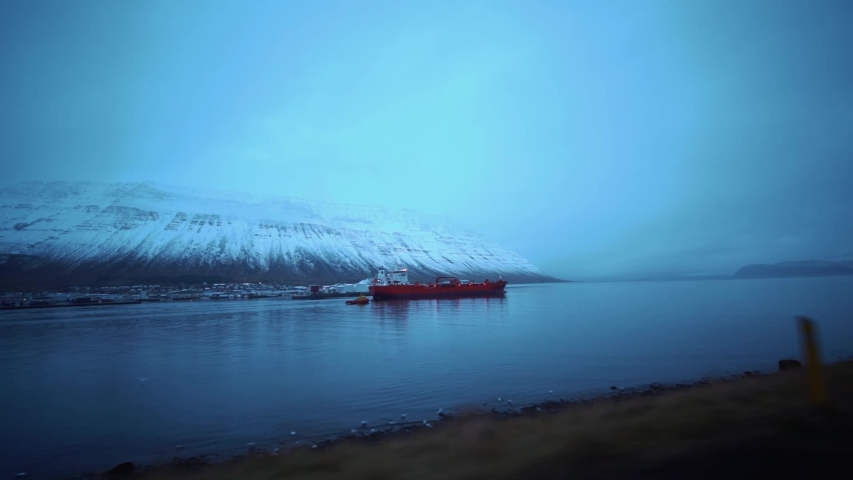 View of massive ice mountain in Island, Reykjavik out of a car window while driving by a red cargo or fishing boat vessel on sea in fjord