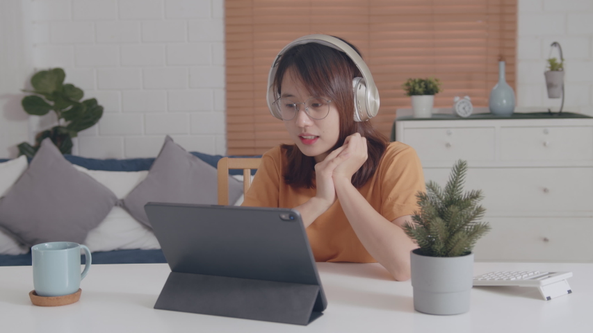 Pretty Young Asian Woman wearing headphones looking at a digital tablet screen Video call learning online on internet, listening and chatting with online teacher or chatting with her friend. Royalty-Free Stock Footage #1047187450