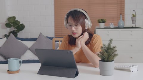 Pretty Young Asian Woman wearing headphones looking at a digital tablet screen Video call learning online on internet, listening and chatting with online teacher or chatting with her friend.