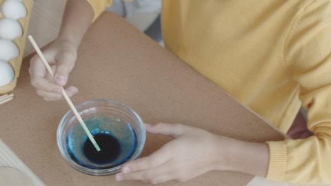 Top view shot of hands of little girl mixing blue food coloring and water in glass bowl while preparing for dying Easter eggs