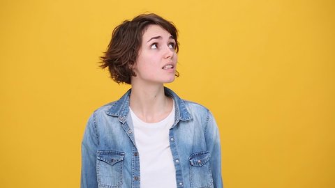 Boring cute brunette young woman in denim jacket white t-shirt posing isolated on yellow background in studio. People lifestyle concept. Looks around, bites her lips, misses and waiting for something