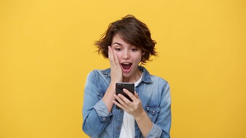 Smiling excited young woman in denim jacket white t-shirt posing isolated on yellow background in studio. People sincere emotions, lifestyle concept. Pointing on cellphone using mobile phone happy win